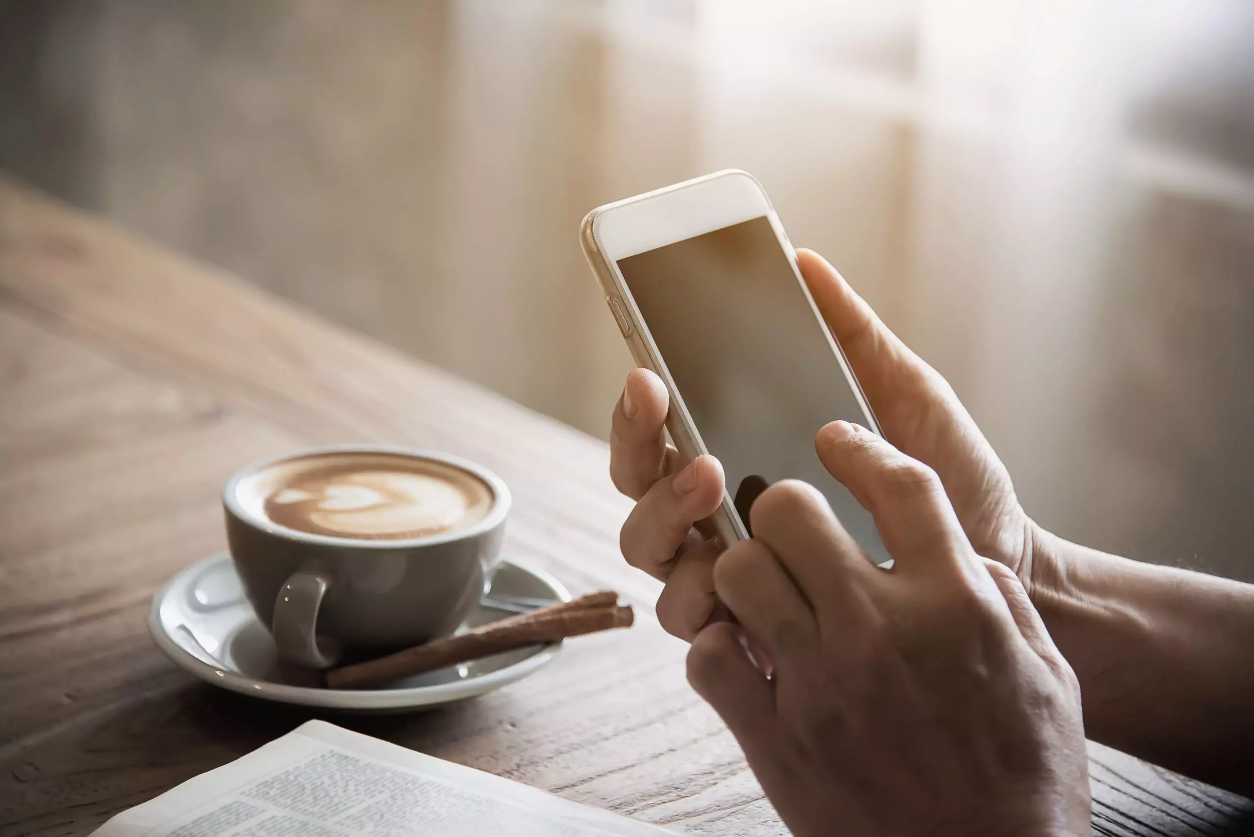 Close das mãos de um homem mexendo em smartphone com um café expresso e um livro na mesa